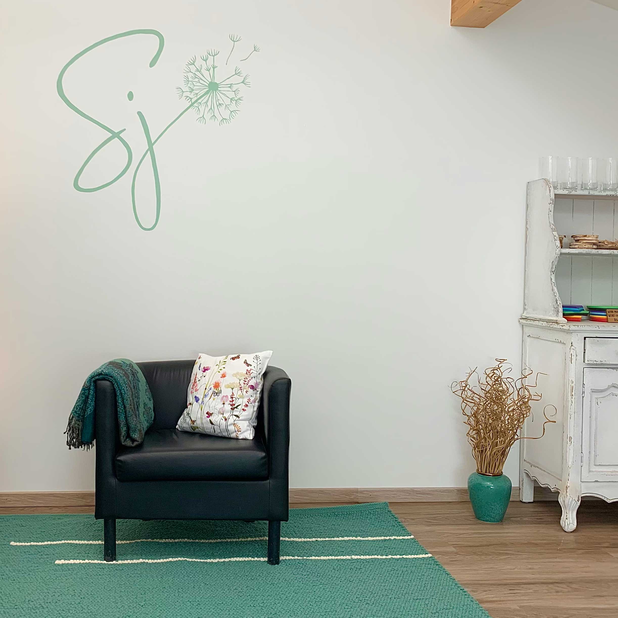 grünes Logo SJ mit Pusteblume an der Wand über Polstersessel mit Decke über der lehne und weißem Polster mit Wiesenblumen, rechst davon am Boden grüne Vase mit zarten Kästchen und Teil einer weißen Kommode mit Gläsern, Holzscheiben und buntem Papier