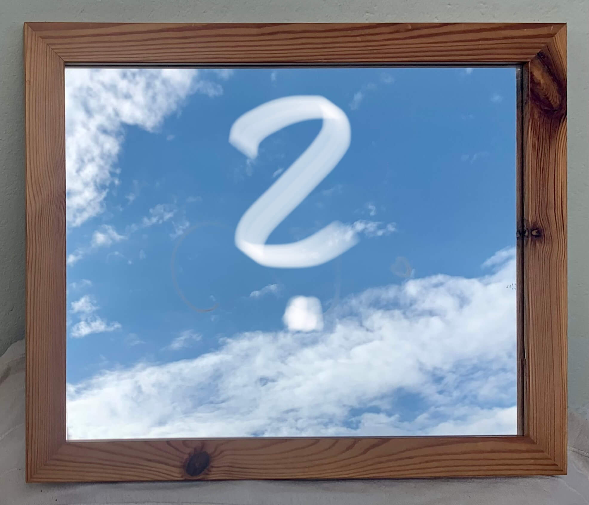 Ein Spiegel mit mittelbraunem Holzrahmen, darin ein großes weißes Fragezeichen vorm blauen Himmel mit weißen Schleierwolken